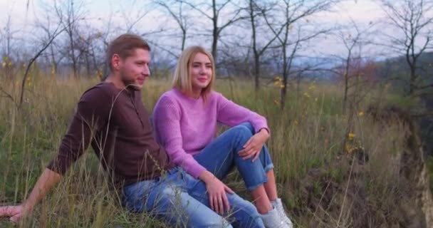 Молода пара туристів сидить на траві в лісі. Стоячи, блондинка дивиться на камеру і посміхається. Біля неї сидить Кавказький молодик.. — стокове відео
