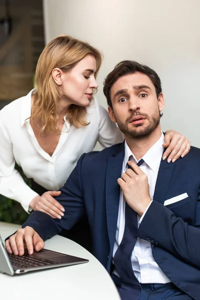 Verführerisch im Bürokonzept. Hübsche Frau berührt männlichen Kollegen, der am Tisch sitzt und verwirrt in die Kamera schaut und seine Krawatte berührt — Stockfoto