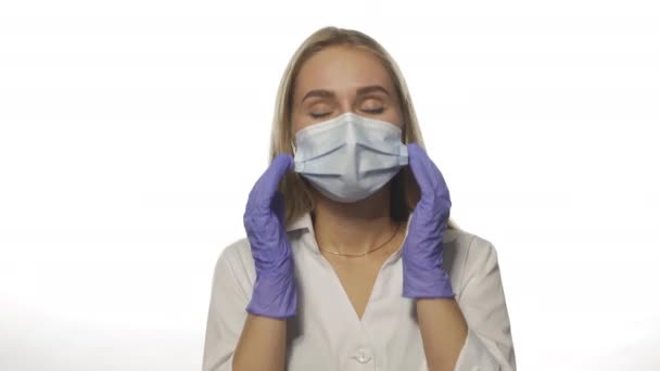Unge læge i en medicinsk maske oplever en migræne på arbejdspladsen, masserer de tidsmæssige lapper i hovedet, ser på kameraet. Isoleret på hvid baggrund. Høj kvalitet 4k opløsning optagelser – Stock-video