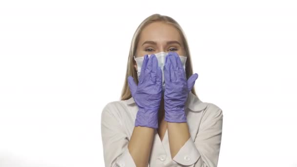 Νέος ξανθός γιατρός που φτιάχνει την ιατρική μάσκα φορώντας μωβ γάντια μιας χρήσης, κοιτάζοντας την κάμερα. Απομονωμένο σε λευκό φόντο. Υψηλής ποιότητας 4k βίντεο ανάλυσης — Αρχείο Βίντεο