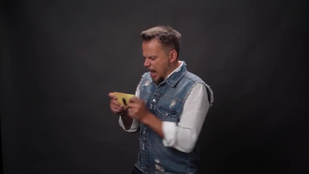 Happy young man playing or vining online game on smartphone or mobile phone using internet or cellular connection wearing denim vest in studio isolated on black background. High quality footage — Stock Video