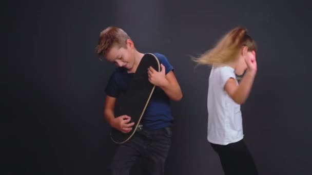 Menino fingindo tocar guitarra usando skate com irmã ou amiga dançando ao lado dele no estúdio isolado em fundo cinza escuro ou preto. Imagens de alta qualidade — Vídeo de Stock