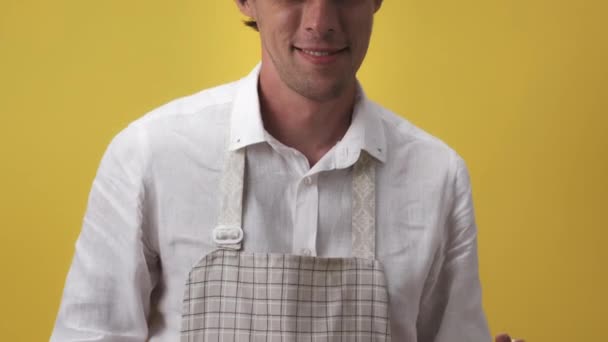 Junger Mann in Schürze drückt Emotionen der Überraschung beim Kochen sehr stark aus. Er trägt ein weißes Hemd auf gelbem Hintergrund. Das Konzept der Emotionen. Kamera rutscht nach oben. Hochwertiges 4k Filmmaterial — Stockvideo