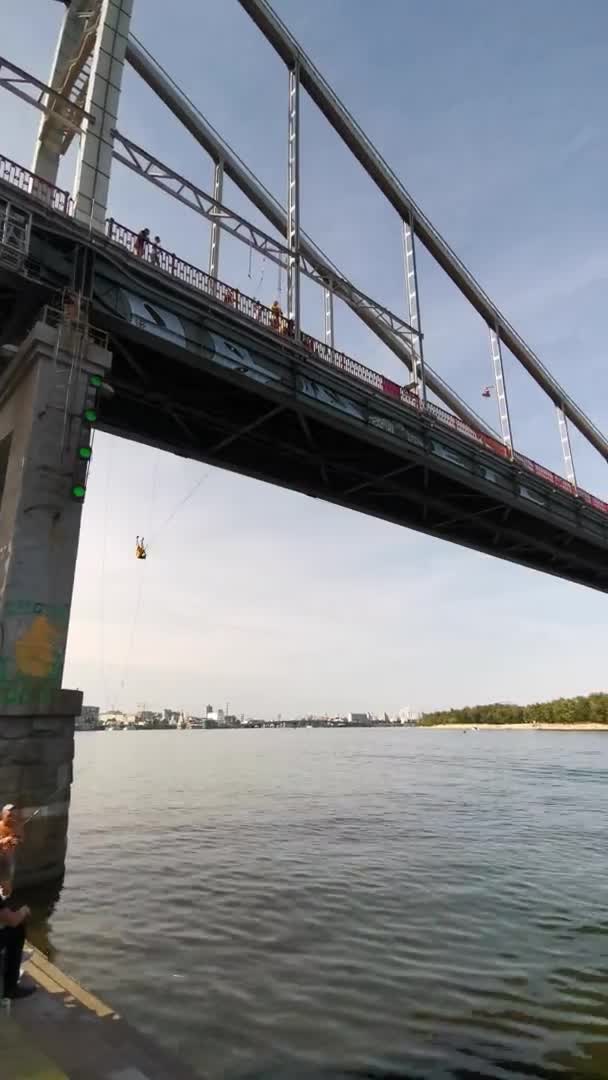 Κάθετη πλάνα του ποταμού Dnipro με πεζόδρομο γέφυρα με θέα τη βιομηχανική πόλη στην οποία σε ένα σχοινί ή μπάντζο swing αρσενικό ή θηλυκό. Υψηλής ποιότητας 4k βίντεο ανάλυσης. 09-06-2020 Κίεβο, Ουκρανία — Αρχείο Βίντεο