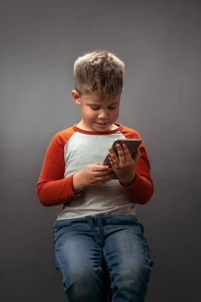 Kırmızı tişörtlü küçük bir çocuk elinde tutan akıllı telefona bakıyor ve gri arka planda izole edilmiş bir şekilde gülümsüyor. İnsani duygular, yüz ifadesi kavramı. Yüz ifadeleri, duygular, duygular — Stok fotoğraf