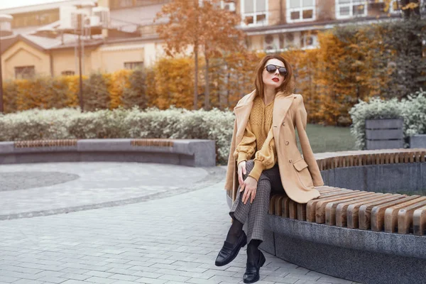 Une belle fille en manteau d'automne et lunettes de soleil beiges s'assoit sur un banc en attendant son rendez-vous ou ses copines. Photo tonique — Photo