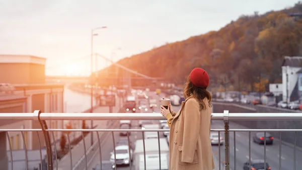 Ung kvinna värmer upp med en kopp kaffe stående baklänges på en gångbro och tittar bort. Porträtt av stilren ung kvinna i höstrock och röd basker utomhus. Tonad bild — Stockfoto