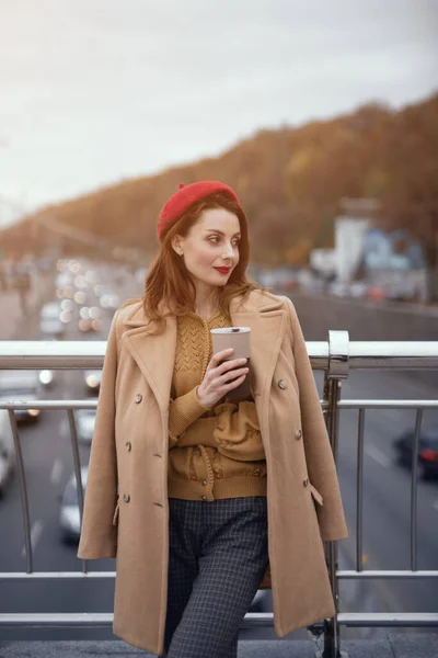 Caddedeki bir yaya köprüsünde duran genç bir kadın kahvesiyle ısınıyor. Sonbahar ceketi ve açık havada kırmızı bereli şık bir genç kadının portresi. Tonu ayarlanmış resim — Stok fotoğraf