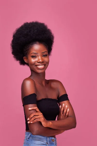 Mooi Afro-Amerikaans meisje met prachtig afro kapsel staan glimlachen met armen gevouwen in zwart top en spijkerbroek geïsoleerd op roze achtergrond — Stockfoto