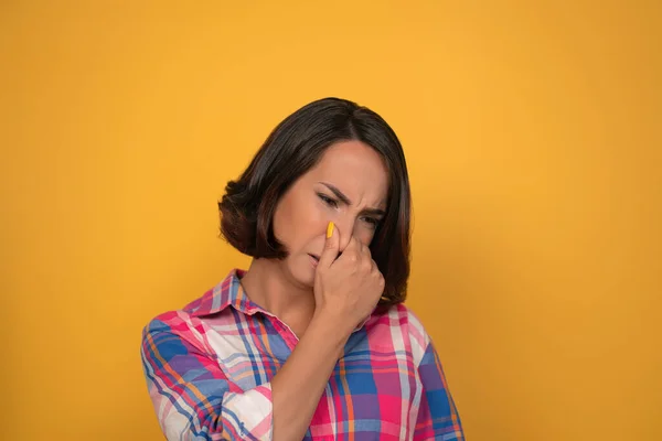 Bad smelling gesture closed nose cute looking woman shows her to her interlocutor, wearing plaid shirt. Isolated on yellow background — Stock Photo, Image