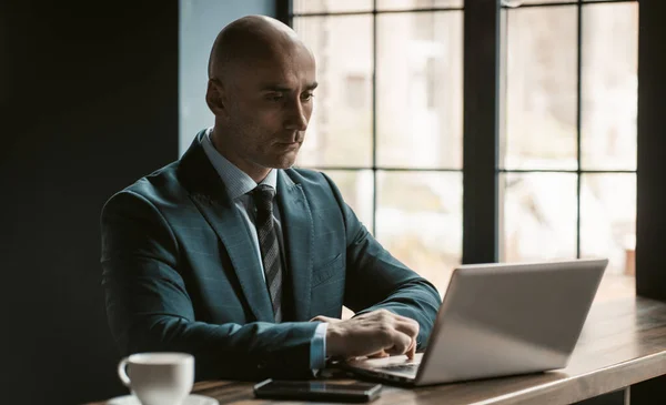 Łysy biznesmen w średnim wieku w apartamencie biznesowym pracujący na laptopie obok otwartych okien w nowoczesnym biurze. Przystojny odważny człowiek pracuje na laptopie w biurze — Zdjęcie stockowe