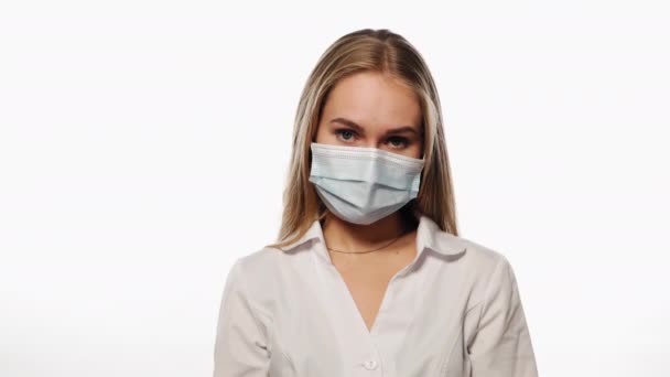 Dunkelblonde Krankenschwester mit medizinischer Maske zeigt Gleichgültigkeit oder Desinteresse oder Apathie mit ihrem Gesicht, blickt in die Kamera. Vereinzelt auf weißem Hintergrund. Hochwertiges 4k-Filmmaterial Stock-Filmmaterial