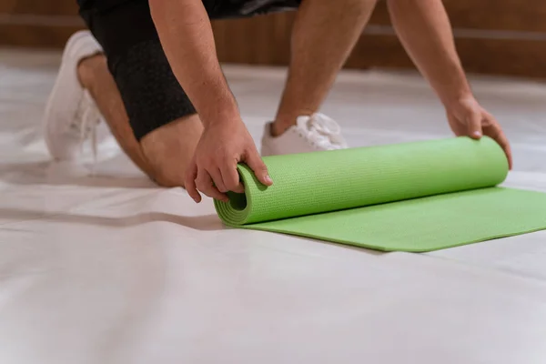 Un joven fuerte y guapo, un atleta, desenrolla una alfombra, preparando un lugar para los deportes. Determinado chico haciendo entrenamiento físico — Foto de Stock