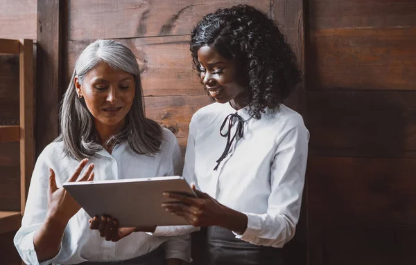 成熟したアジアの女性と若いアフリカのビジネス女性が一緒に木製のオフィスの壁の横にあるデジタルタブレットを保持いくつかの財務的な側面について話して立っている — ストック写真