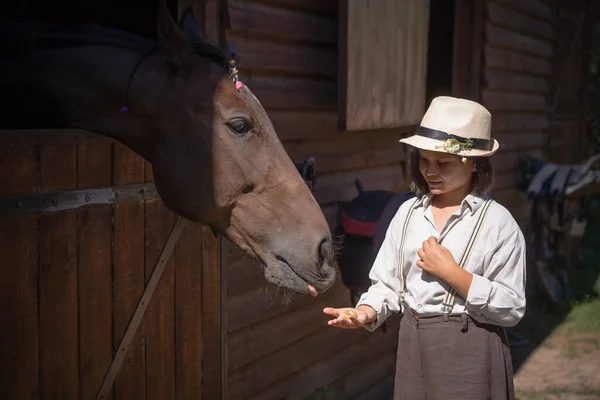 Charmante jeune fille en vêtements vintage debout avec son cheval brun qui sort tête de la grange pendant la journée ensoleillée. Concept d'amitié — Photo