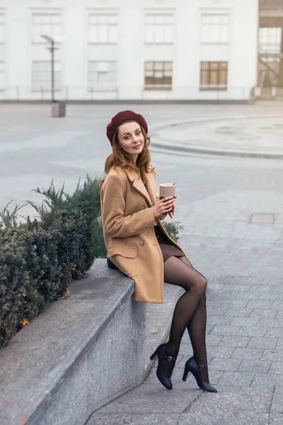 Hermosa chica en un abrigo de otoño y beige se sienta en un macizo de flores de mármol esperando a su cita o novias. Foto tonificada — Foto de Stock