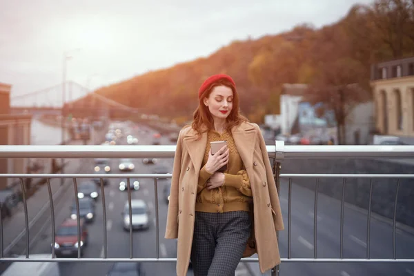 Porträtt av stilren ung kvinna i höstrock och röd basker utomhus. Tender ung kvinna värma upp med kopp kaffe på gatan kvinnligt mode stående på en gångbro. Tonad bild — Stockfoto