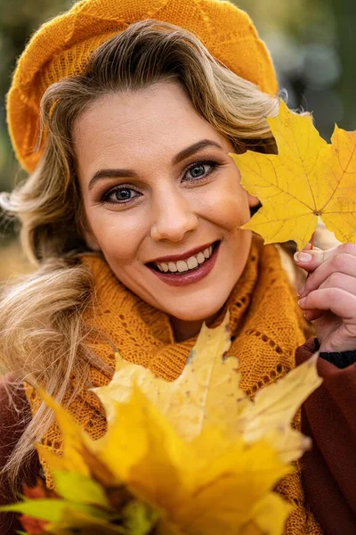 Aus nächster Nähe. Junge Frau in beigefarbener Strickmütze mit Herbstblättern in der Hand und herbstgelbem Garten oder Park. Schöne lächelnde junge Frau im Herbstlaub — Stockfoto