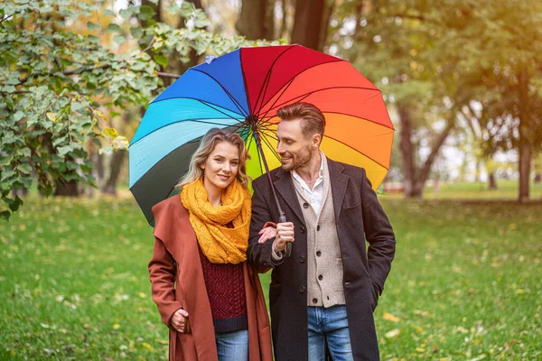 Ein schönes junges Paar spaziert unter einem regenbogenfarbenen Regenschirm durch den Park. Ein schönes Mädchen geht Arm in Arm mit einem Mann bei Regenwetter durch den herbstlichen Park — Stockfoto