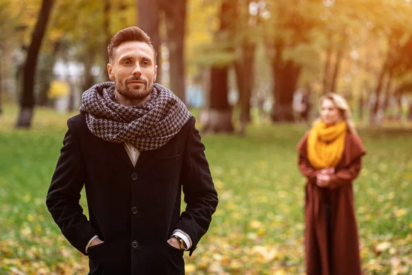 Mannen står i en rock och en scarf i parken och väntar på sin flickvän eller fru som står bakom honom eller ser ut som honom bakifrån. Unga par utomhus i en park på en vacker höstdag — Stockfoto