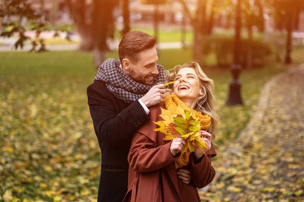 Jóvenes felices enamorados, hombre abrazando a la mujer por detrás acariciando su mejilla, feliz pareja caminando en un parque de otoño con elegantes abrigos y recogiendo hojas caídas. Concepto de familia y personas — Foto de Stock