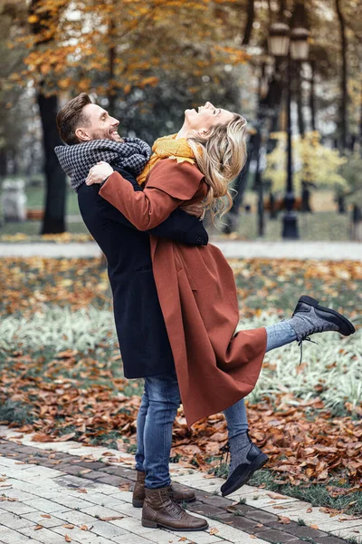 Man lyfter en flicka kramas skrattar i höstparken med en söt kram. Utomhus bild av ett ungt par kär har roligt att ha en kram i en höstpark. Närbild full längd.tonad bild — Stockfoto