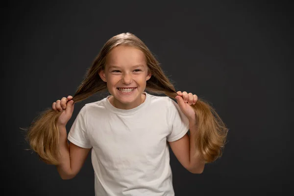 Szczęśliwy uśmiech i trzymając dwa ogony kucyka 8,10 lat dziewczyna trzyma jej włosy w białej koszuli uśmiecha się trochę uśmiecha się do kamery odizolowany na ciemnoszarym lub czarnym tle — Zdjęcie stockowe
