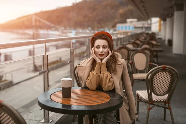 Hayal kırıklığına uğramış ya da hayal kırıklığına uğramış genç bir Fransız kadın kahve fincanı kadın modeliyle açık hava restoranının terasında oturuyor. Sonbahar ceketi ve açık havada kırmızı bereli şık bir genç kadının portresi. — Stok fotoğraf