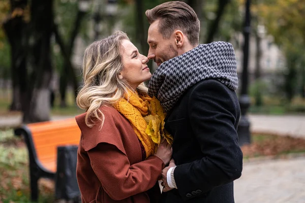 Apasionado pareja guapo chico sostener sus novias manos y tocar la nariz de pie en el parque de la ciudad de otoño con abrigos y bufandas. Pareja sonriente abrazándose en el parque de otoño — Foto de Stock