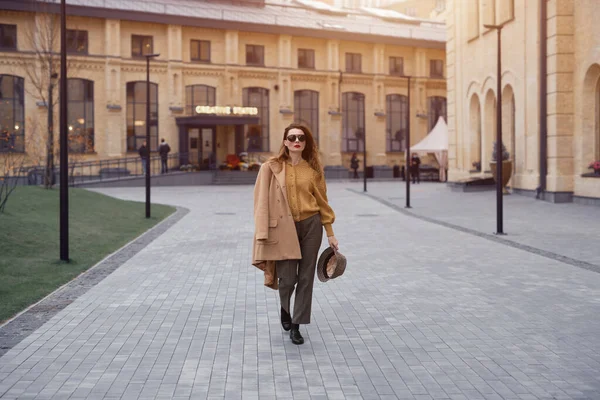 Mode jeune femme en lunettes de soleil et accroché sur une épaule automne manteau beige marchant dans la rue posant joyeusement pour l'appareil photo. Photo teintée — Photo