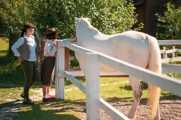 Glad bondgård. Mor och dotter i vintagekläder står nära hagen med en vacker vit häst. Flickan smeker en häst. Familjen på ranchen — Stockfoto