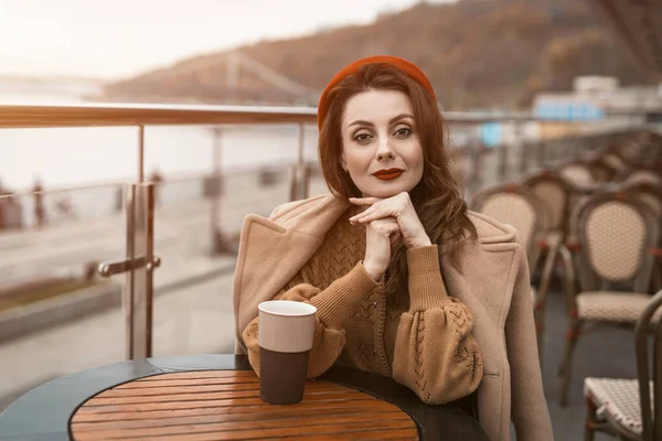 Lokanta terasında elinde kahve fincanıyla kameraya bakan güzel bir Fransız genç kadın. Sonbahar ceketi ve açık havada kırmızı bereli şık bir genç kadının portresi. — Stok fotoğraf