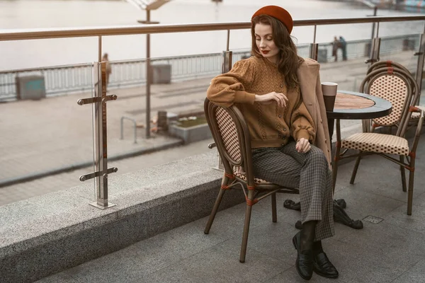 Attendre ou frustré jeune femme française assise à l'extérieur restaurant de rue à l'air frustré vers le bas. Portrait de la jeune femme élégante portant manteau d'automne et béret rouge à l'extérieur — Photo