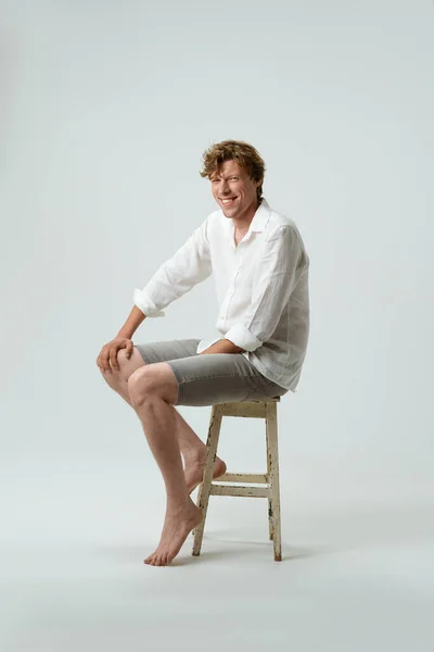 Volledige lengte van een zitten op stoel lachende jongeman kijken naar camera geïsoleerd op witte achtergrond. Gezichtsuitdrukkingen, emoties, gevoelens — Stockfoto
