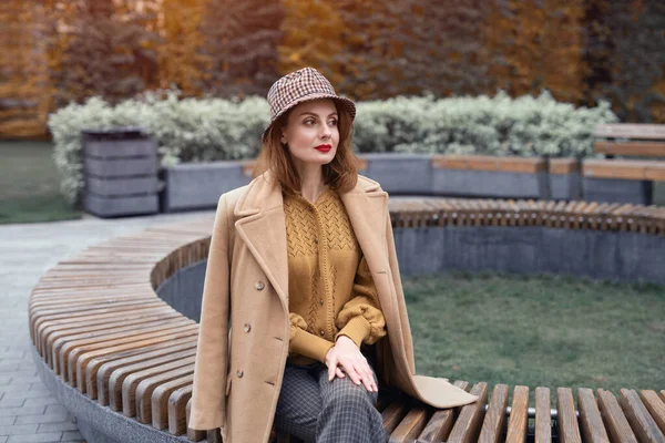 Charmante jeune femme en manteau beige et chapeau à carreaux s'assoit sur un banc rond en attendant son rendez-vous ou ses copines. Concept de marche d'automne. Photo teintée — Photo