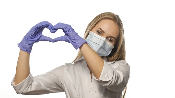 Bela jovem enfermeira de roupão branco e máscara médica mostra sinal de amor com as mãos levantando-se, olhando para a câmera. Isolado sobre fundo branco — Fotografia de Stock
