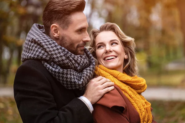 Κλείσε. Ευτυχισμένοι στην αγάπη νέοι άνθρωποι, άντρας αγκαλιάζει γυναίκα από πίσω όταν τον κοιτάζει, ευτυχισμένο ζευγάρι που περπατά σε ένα πάρκο το φθινόπωρο φορώντας καλαίσθητα παλτά και μαζεύοντας πεσμένα φύλλα — Φωτογραφία Αρχείου