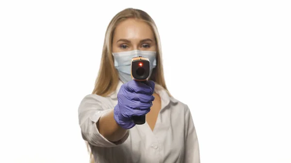 Медична медсестра в захисній масці вказує термометр датчика на камеру в білому медичному одязі лікарні. Молода блондинка-волохата медсестра перевіряє пацієнтів або колегу температури. 4k кадри — стокове фото