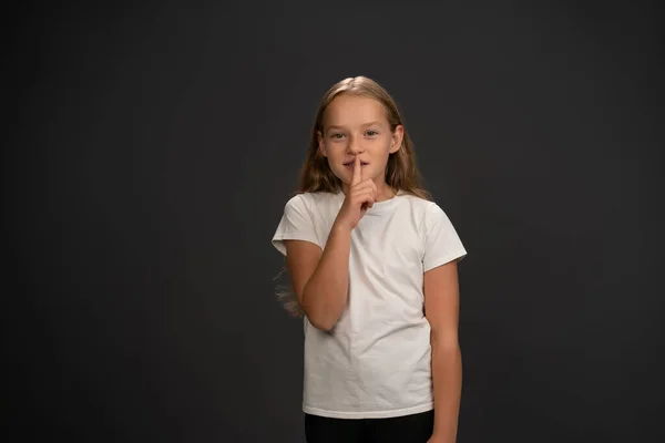 Little girl holding her finger on her lips telling everyone to keep silence wearing white t shirt smiling at the camera isolated on dark grey or black background — Stock Photo, Image