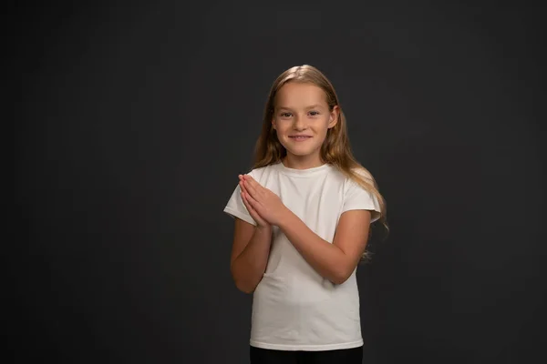 Jubelndes kleines Mädchen, das mit zusammengelegten Händen glücklich in die Kamera blickt und weißes T-Shirt auf dunkelgrauem oder schwarzem Hintergrund trägt. — Stockfoto