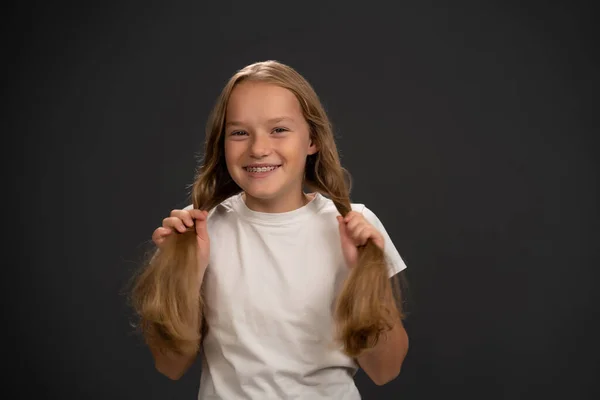 Sorrindo segurando duas caudas de pônei menina vestindo t-shirt branca sorrindo um pouco sorrindo para a câmera isolada no fundo cinza escuro ou preto — Fotografia de Stock