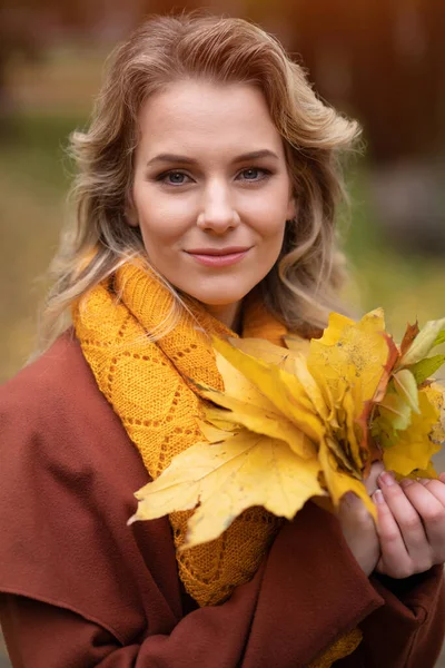 Красива молода жінка позує на камеру з осіннім листям в руках і падає жовтим садом або парком. Красива усміхнена молода жінка в осінньому листі — стокове фото