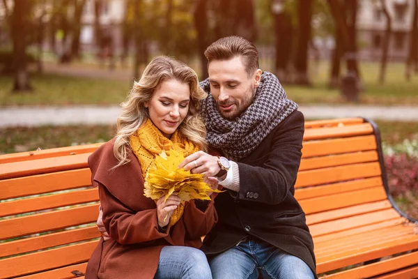 Abrazando pareja feliz sentada suavemente romántica abrazada en un banco en el parque usando abrigos y bufandas Recogiendo un ramo de hojas caídas. Concepto de historia de amor — Foto de Stock