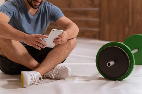 No hay disparo en la cara. Joven buscando en línea o mensajes de texto sentado en un piso con teléfono inteligente, barra de fitness de tono negro y verde, equipo para el concepto de entrenamiento con pesas. concepto de estilo de vida saludable — Foto de Stock