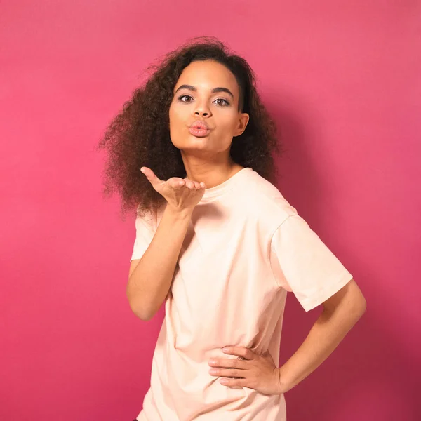 Linda jovem afro-americana com cabelo afro enviando beijos de ar, olhando positivamente para a câmera vestindo t-shirt de pêssego isolado no fundo rosa. Conceito de beleza — Fotografia de Stock