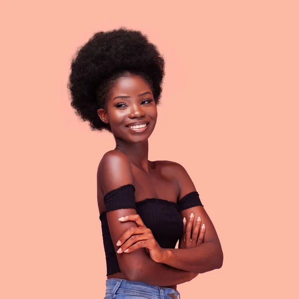 Square portrait of happy charming African American girl with amazing afro hairstyle smiling with arms folded. Dark-skinned lady wearing black bare shoulders top and denim jeans on peach background — Stock Photo, Image