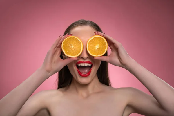Engraçado bela beleza modelo jovem mulher segurando duas fatias de laranja suculentas como seus olhos ou óculos. Encantadora senhora engraçada alegre com lábios vermelhos e cabelos longos isolados no fundo rosa — Fotografia de Stock