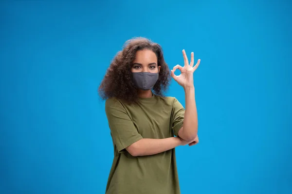 Menina afro-americana jovem em cáqui t-shirt em máscara facial reutilizável mostrando gesto OK, para evitar que os outros de corona COVID-19 e SARS cov 2 infecção isolada em fundo rosa — Fotografia de Stock