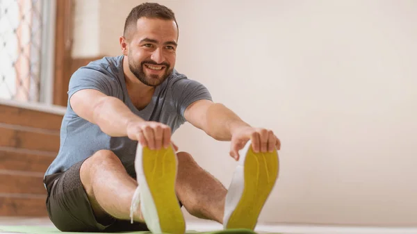 Happy smiling athletic young man stretching out training at home. Amateur sports man in workout activity sitting on pad and stretch leaning to his toes. Hardworking guy exercising in apartment — Stockfoto