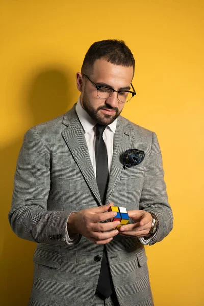 Młody przystojny mężczyzna w okularach odziany w apartament rozwiązujący zagadkę w dłoniach z kostki kieszonkowej 2x2x2, patrząc na nią z góry. Izolacja na żółtym tle — Zdjęcie stockowe